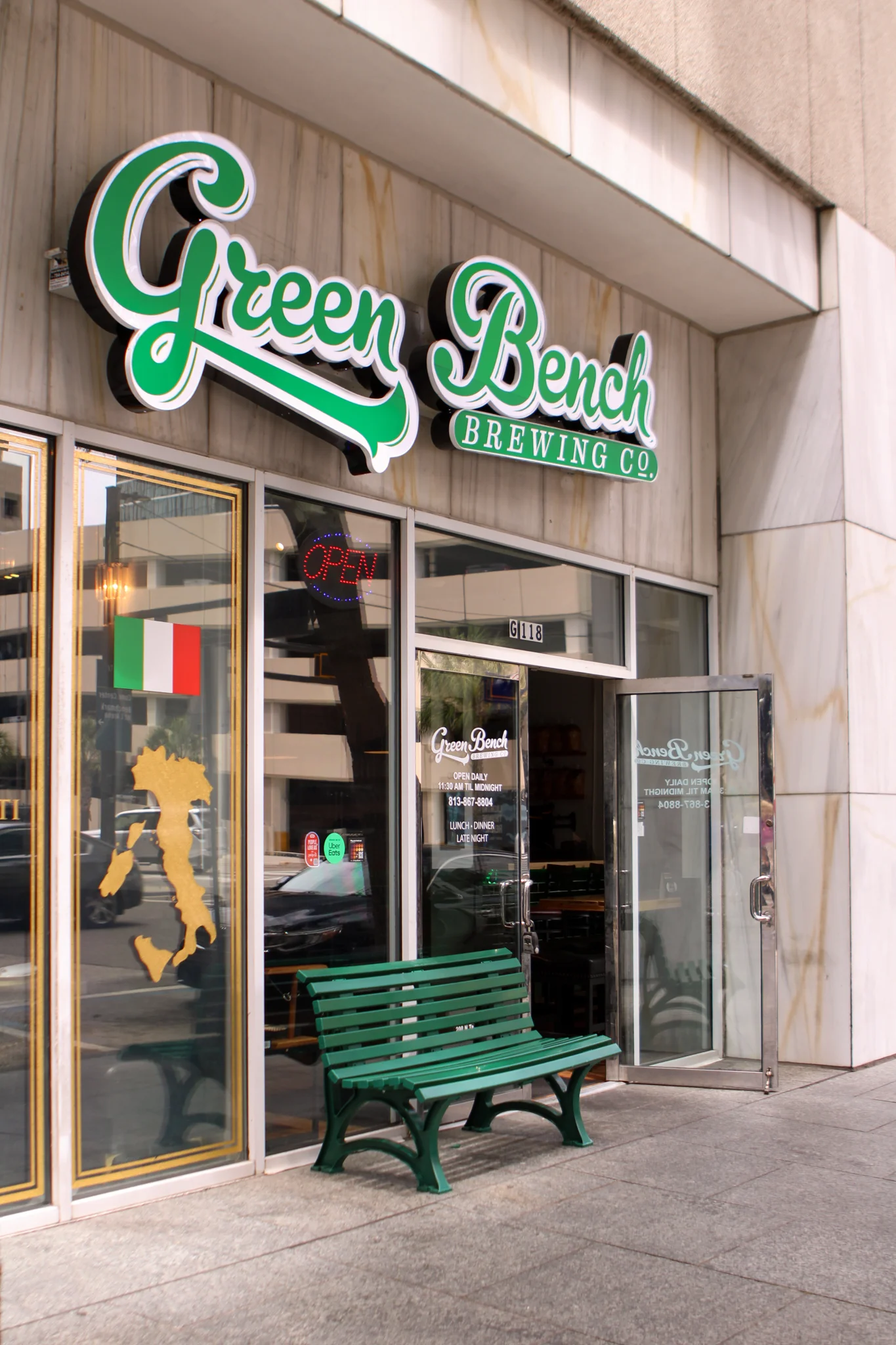 The entrance of a brewing company featuring a green sign and a small green bench outside. The glass door displays an open sign and information about opening hours.
