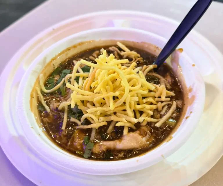A bowl of spicy soup topped with shredded cheese and garnished with cilantro, served in a white disposable container.