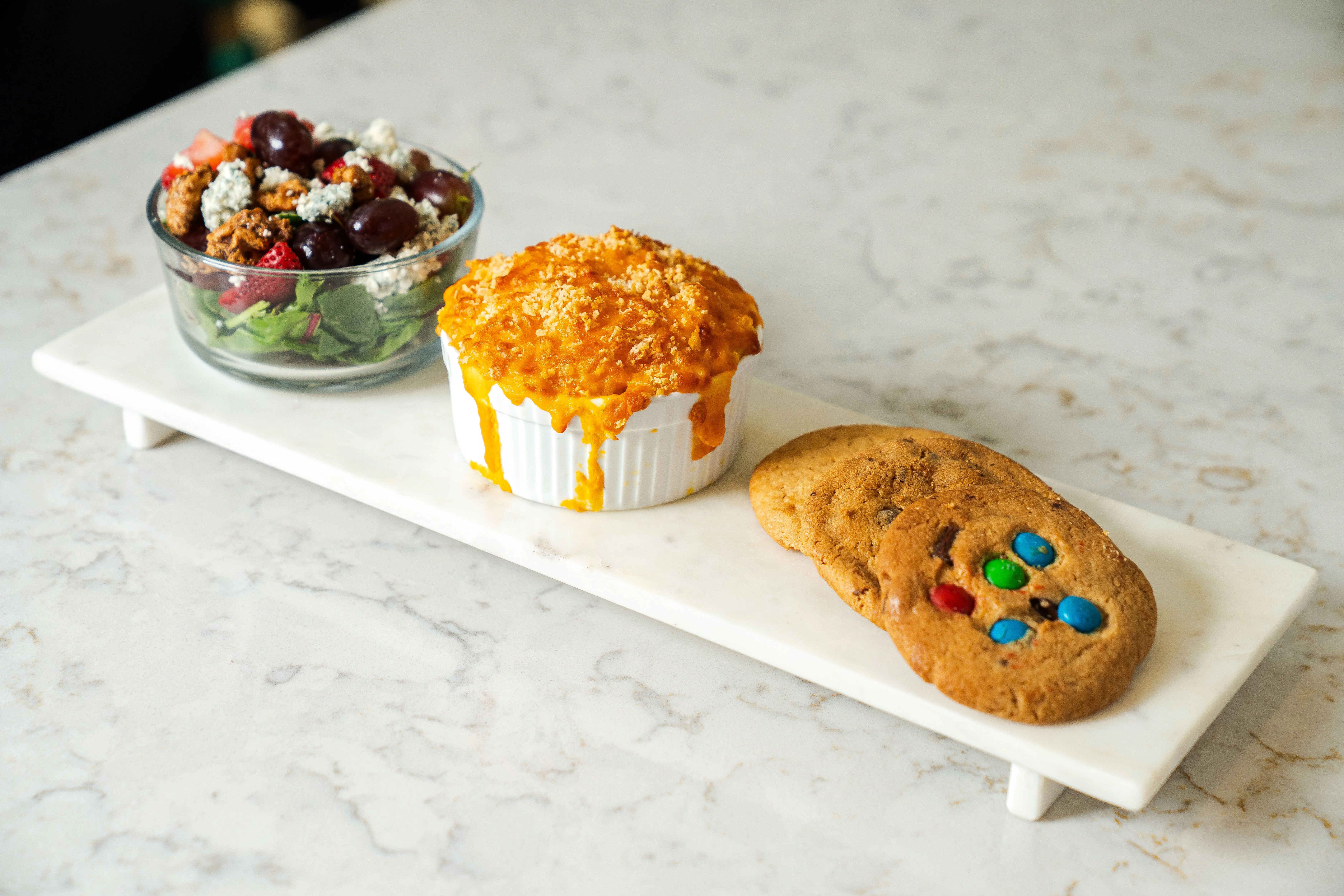 A serving of mac and cheese alongside a fresh salad and two colorful cookies on a marble tray.