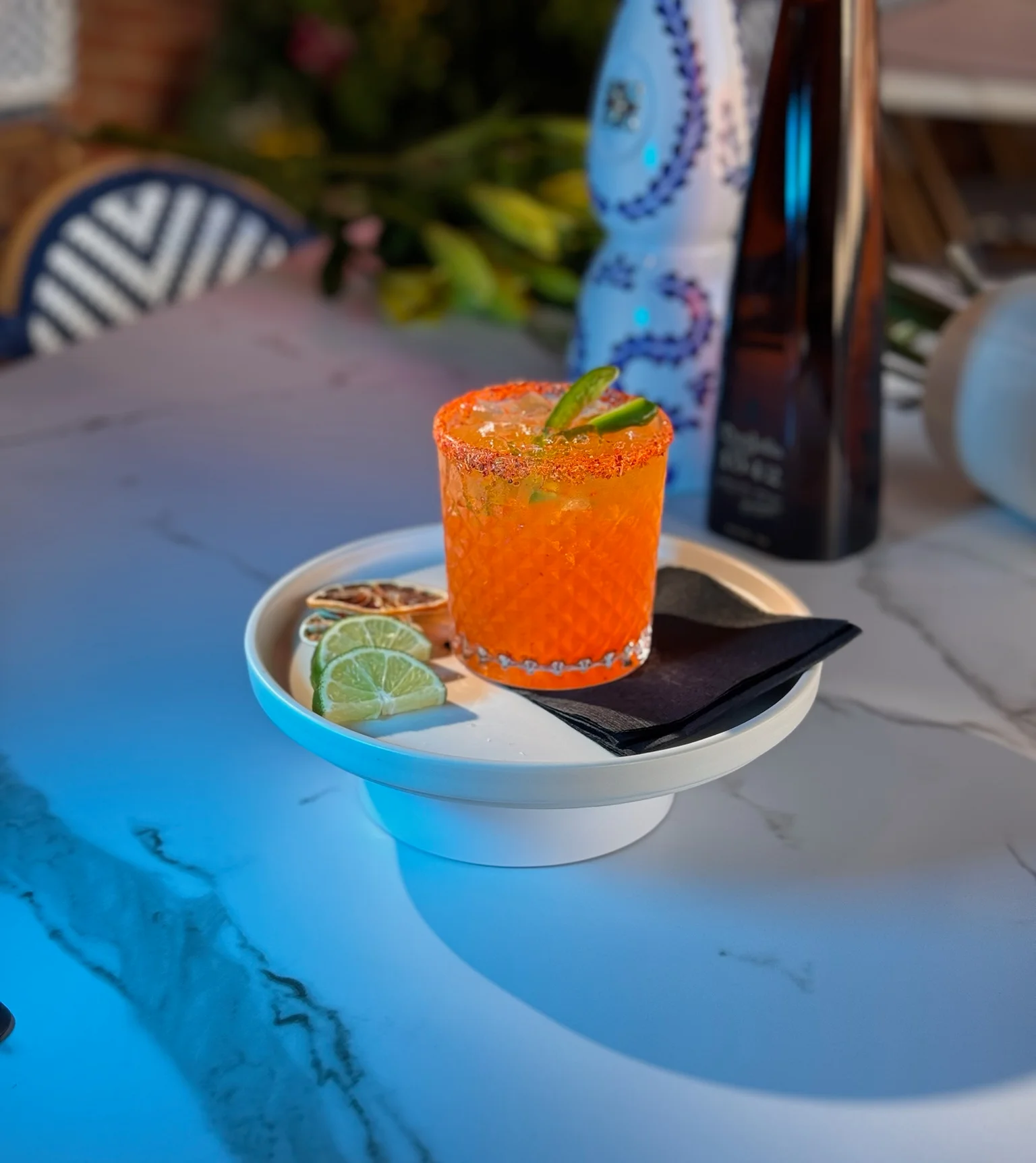 A vibrant orange cocktail served in a decorative glass on a white plate. Fresh lime slices and a black napkin accompany the drink.