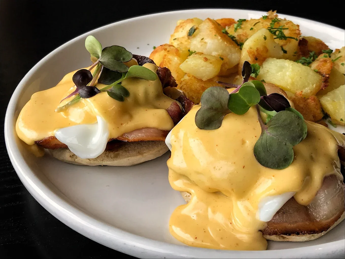 A plate featuring two eggs Benedict with ham and a creamy sauce, accompanied by crispy golden potatoes.