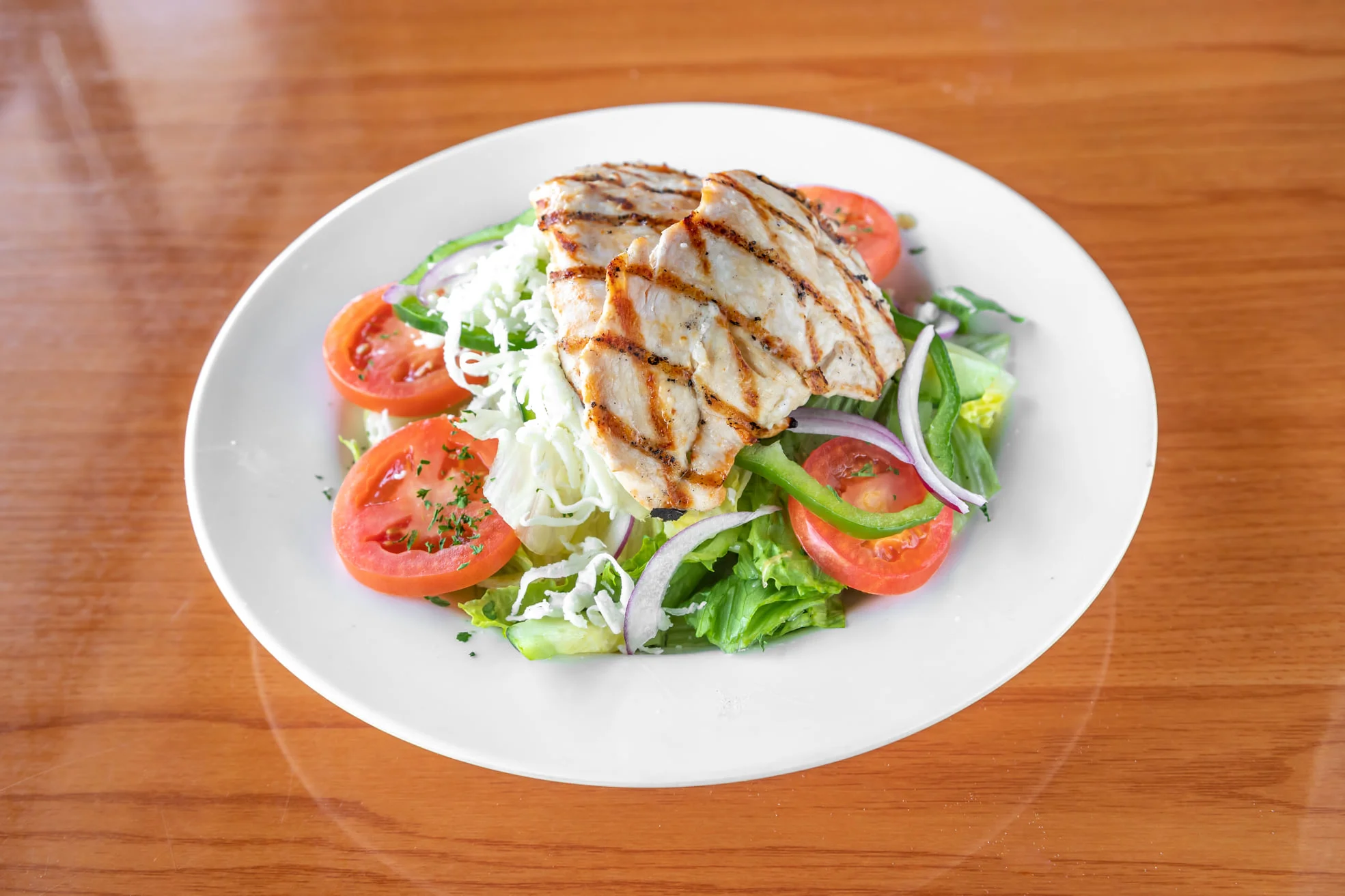 A plate of grilled chicken atop a fresh salad with tomatoes, lettuce, and green peppers, garnished with herbs.