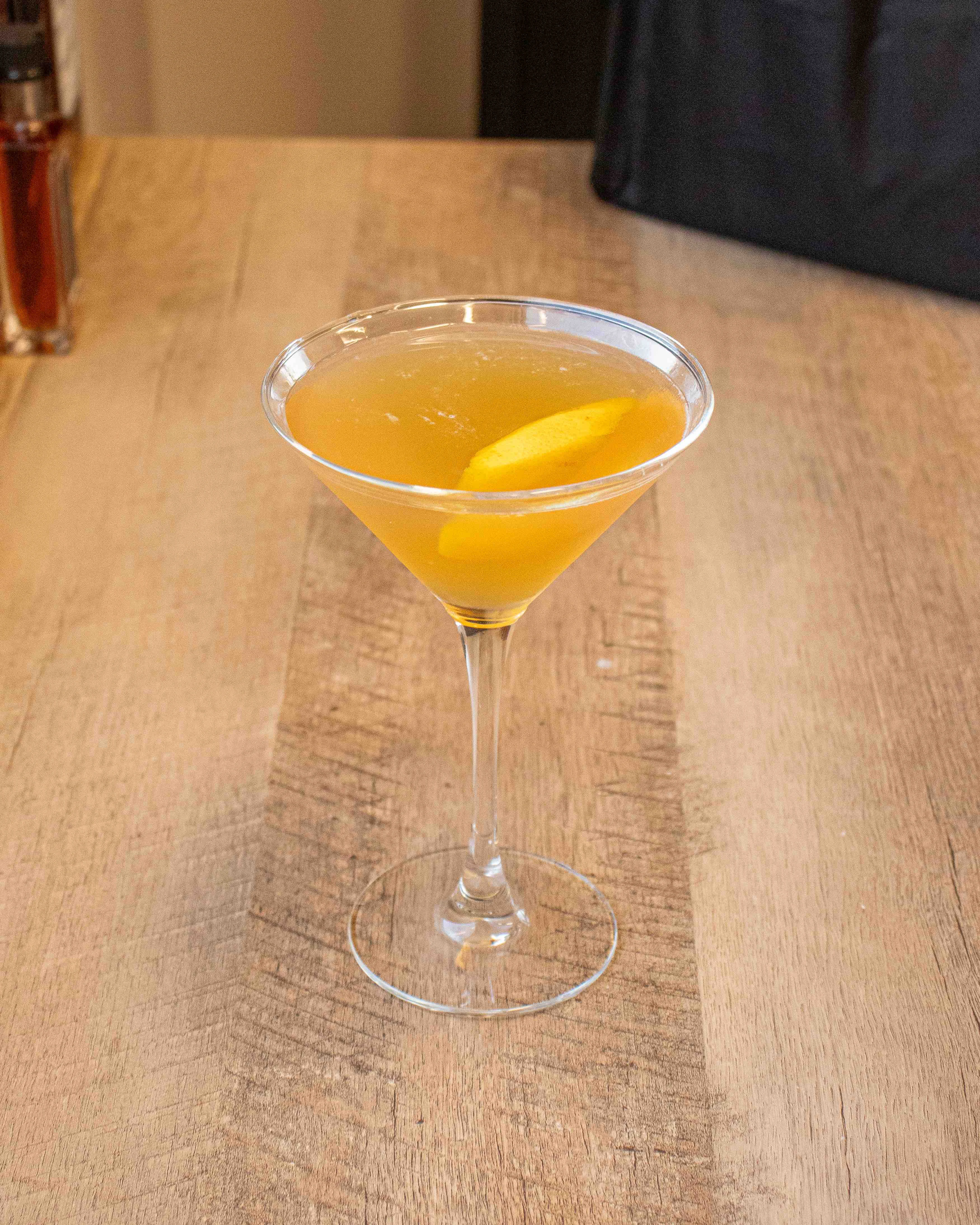 Cinnamon Peach Martini sits on a wooden table with an orange twist garnish. 