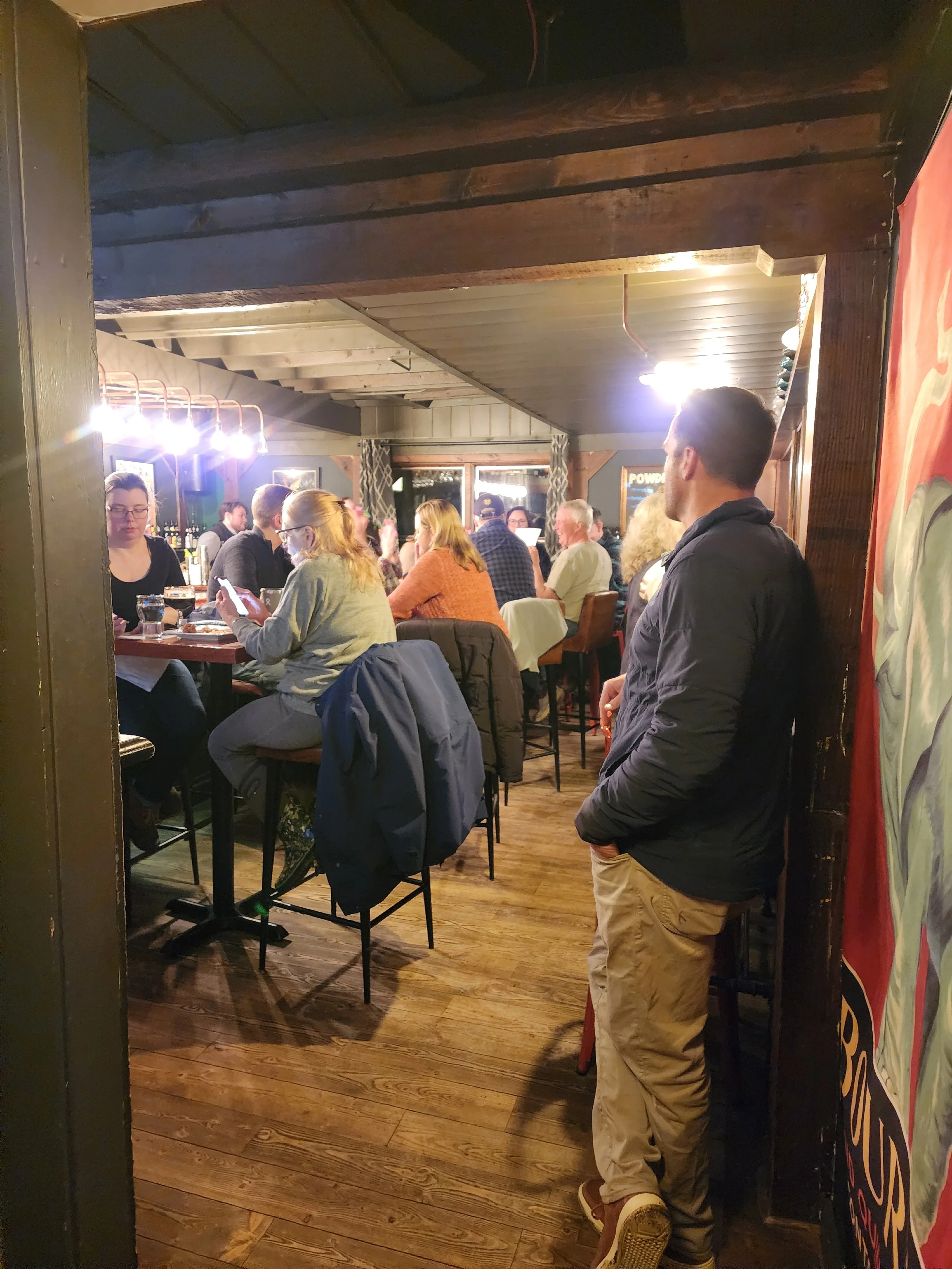 Busy restaurant dining room with guests, exposed wood beams, and warm lighting