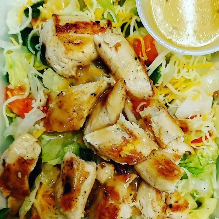 A chicken salad topped with grilled chicken pieces, cheese, and mixed vegetables, served with a dressing on the side.