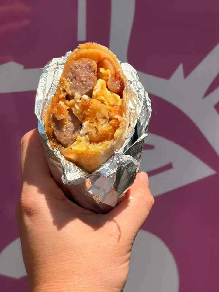 A breakfast burrito filled with sausage, eggs, and crispy ingredients, wrapped in foil and held by a hand.