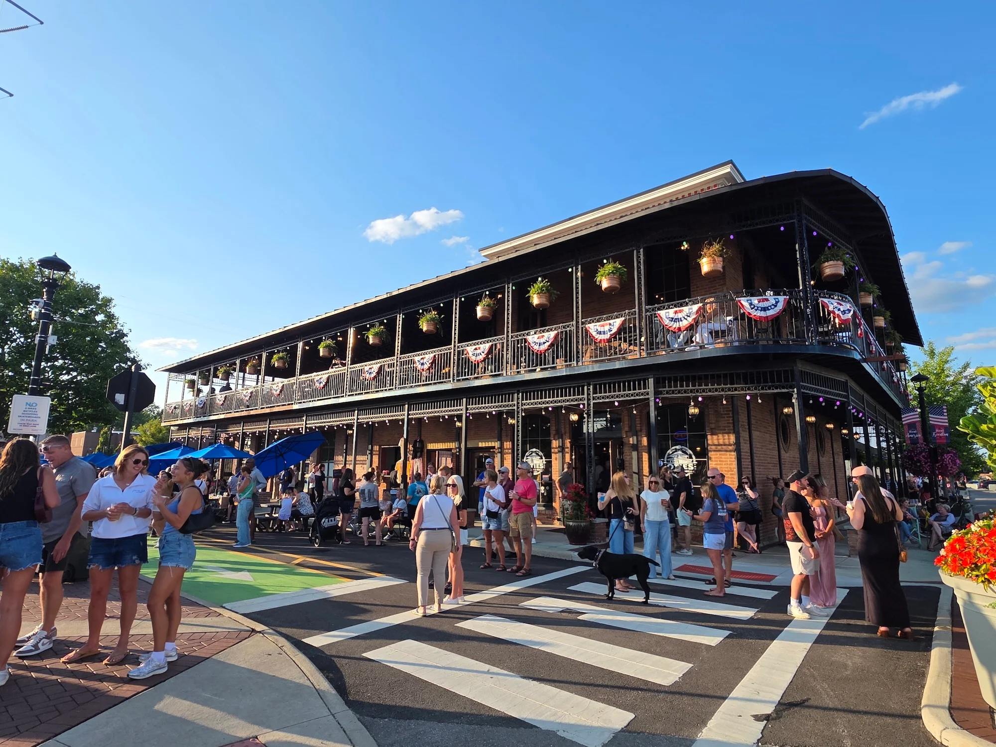 A bustling outdoor area with a two-story building adorned with hanging plants and bunting. Pedestrians enjoy the lively atmosphere on a sunny day.