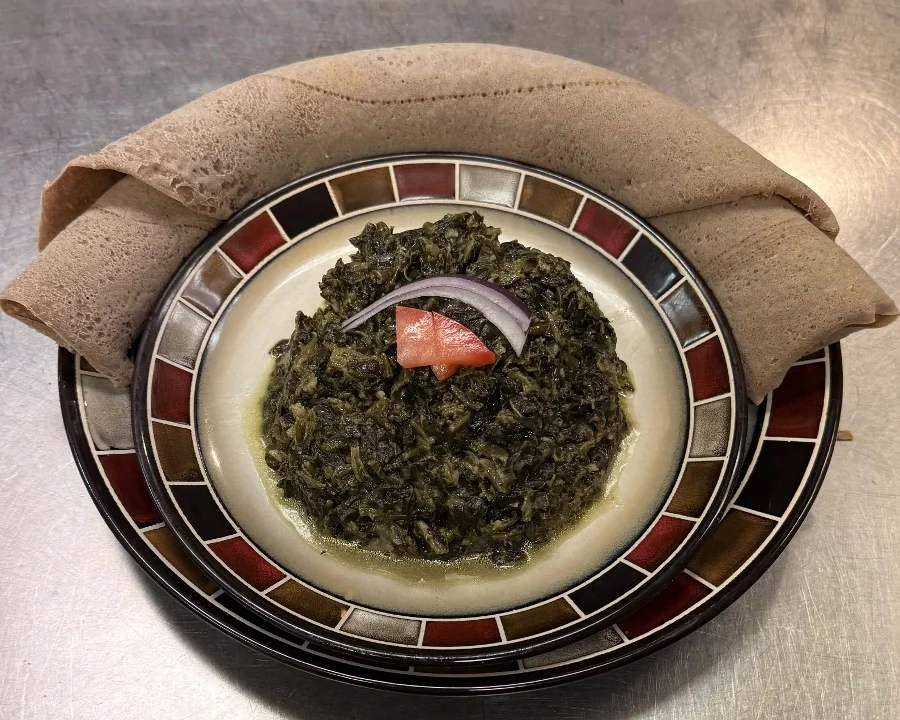A serving of sautéed greens topped with slices of tomato and onion, served with flatbreads on a decorative plate.