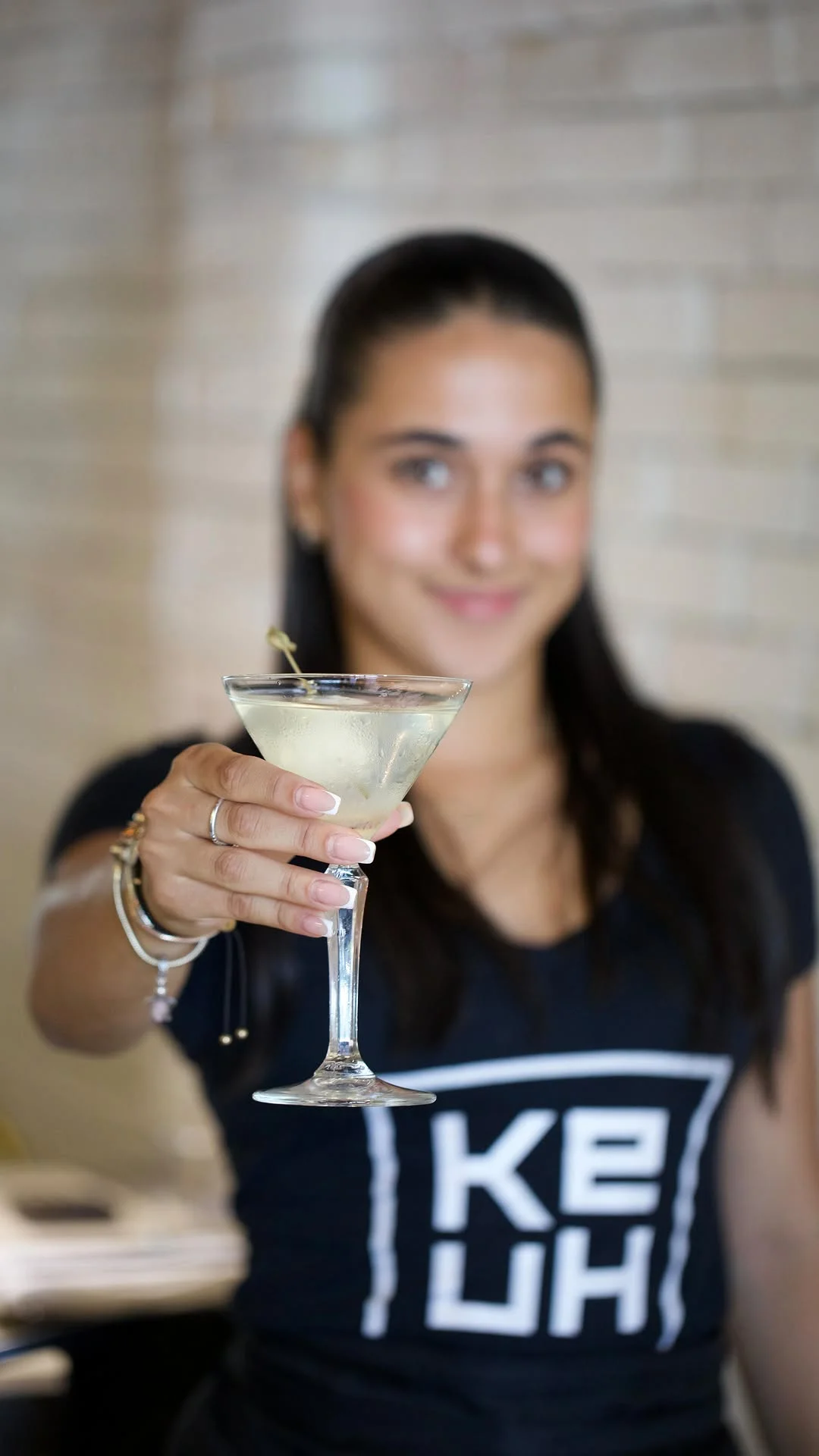 A server holding out a chilled martini cocktail in a stem glass, with the Ke-uH's logo visible on her black uniform