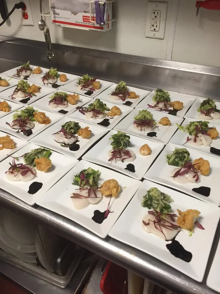 A row of elegantly arranged plates featuring various ingredients including greens, fried items, and sauces, all presented in a professional kitchen setting.