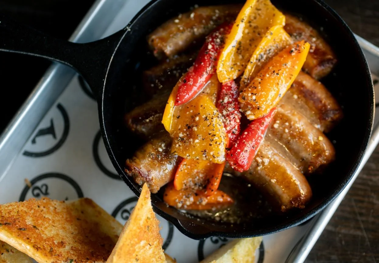 A cast-iron skillet filled with sausages and colorful bell peppers, seasoned and surrounded by toasted bread.