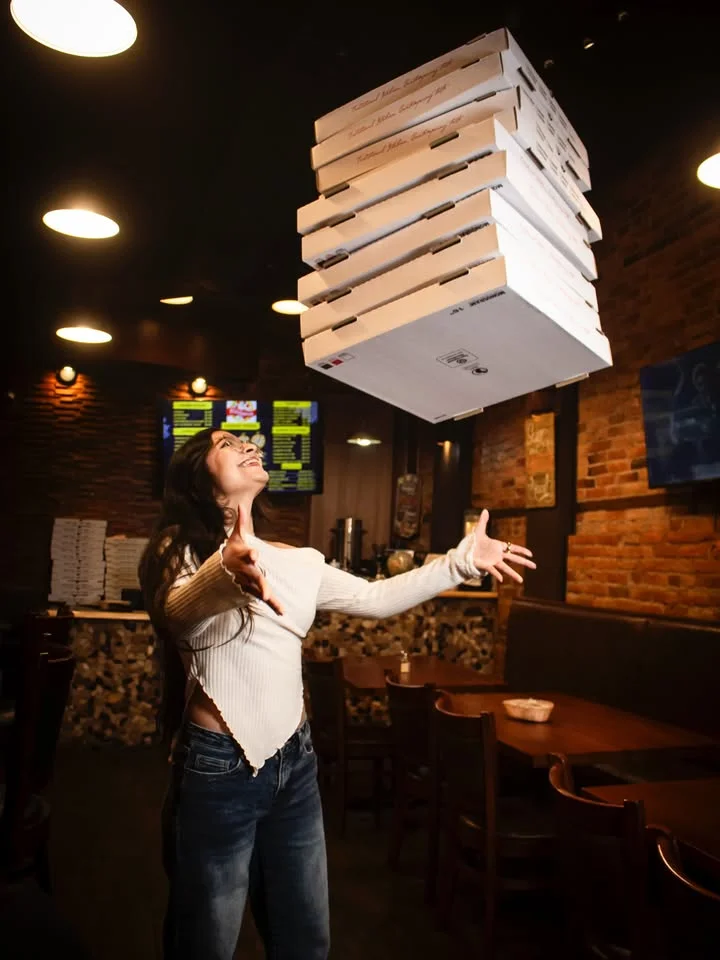 A person joyfully tossing pizza boxes in the air in a lively dining environment with brick walls.