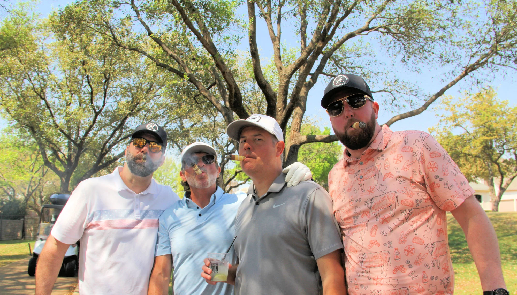 Friends enjoying cigars and drinks outdoors beneath large oak trees