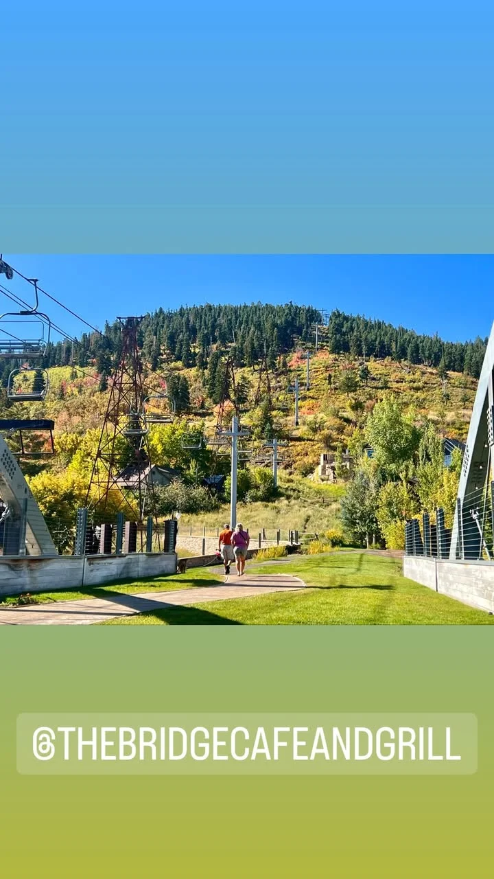 A scenic view of a walking path with two people strolling near a ski lift and vibrant autumn foliage in the background.