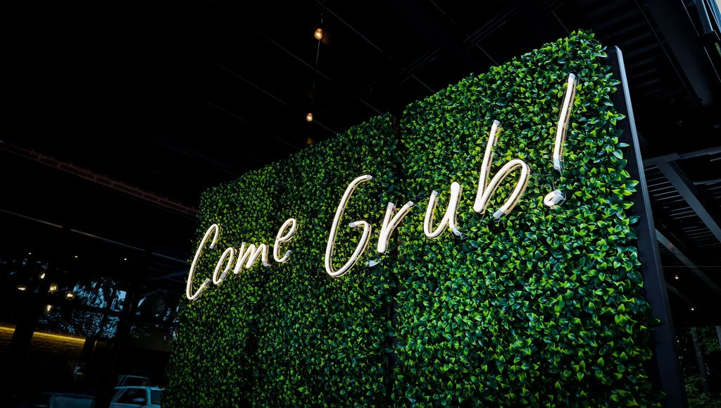 Restaurant signage with lush greenery saying 'Come Grub!'