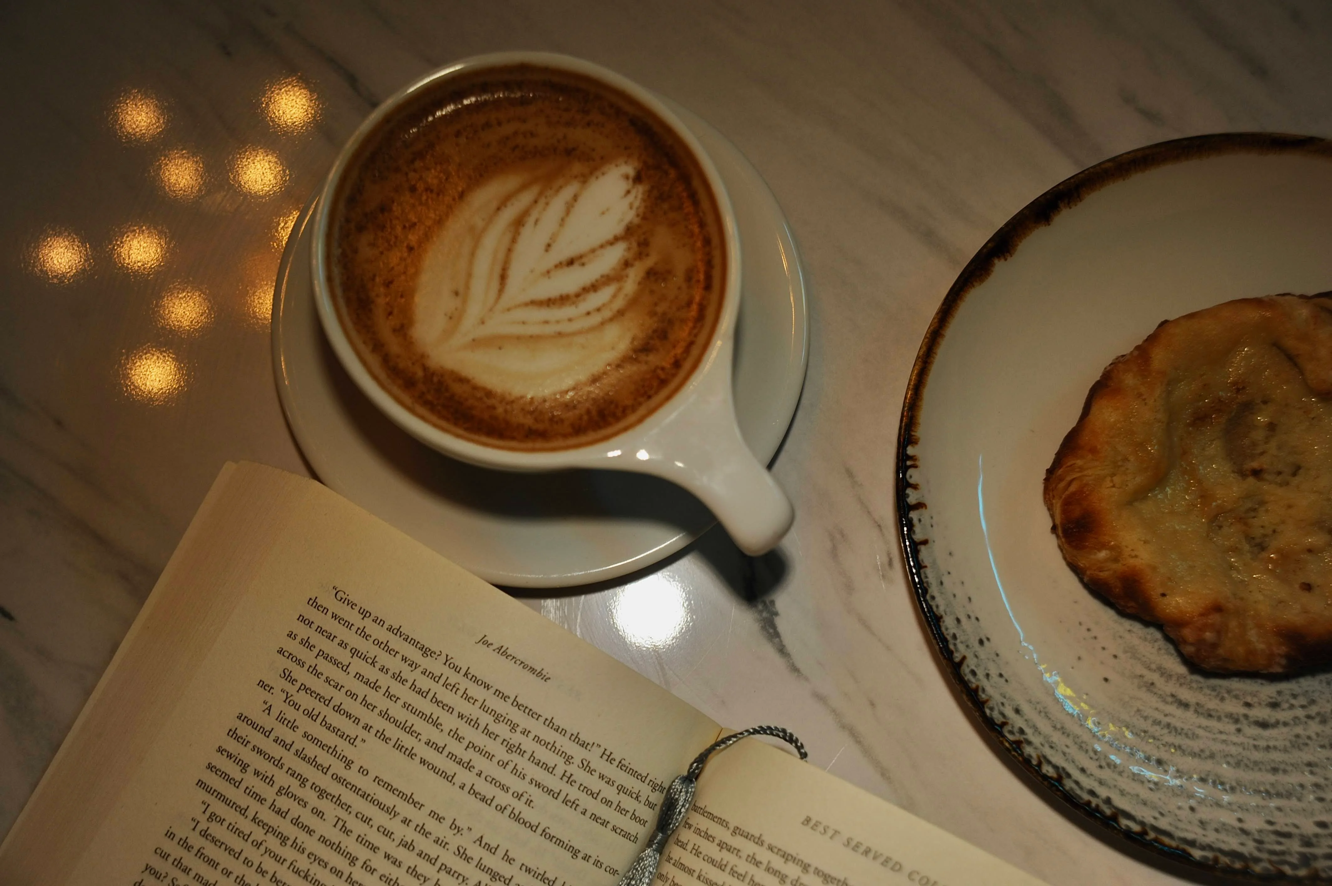 A cozy scene featuring a latte with leaf art, a pastry on a plate, and an open book on a marble surface.