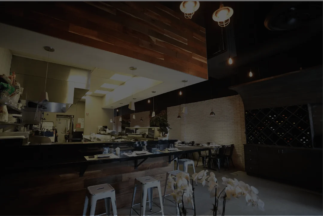 Ke-uH sushi bar featuring a black countertop with formal place settings, silver industrial stools, and a wooden base, set against a white brick wall and a professional kitchen area.