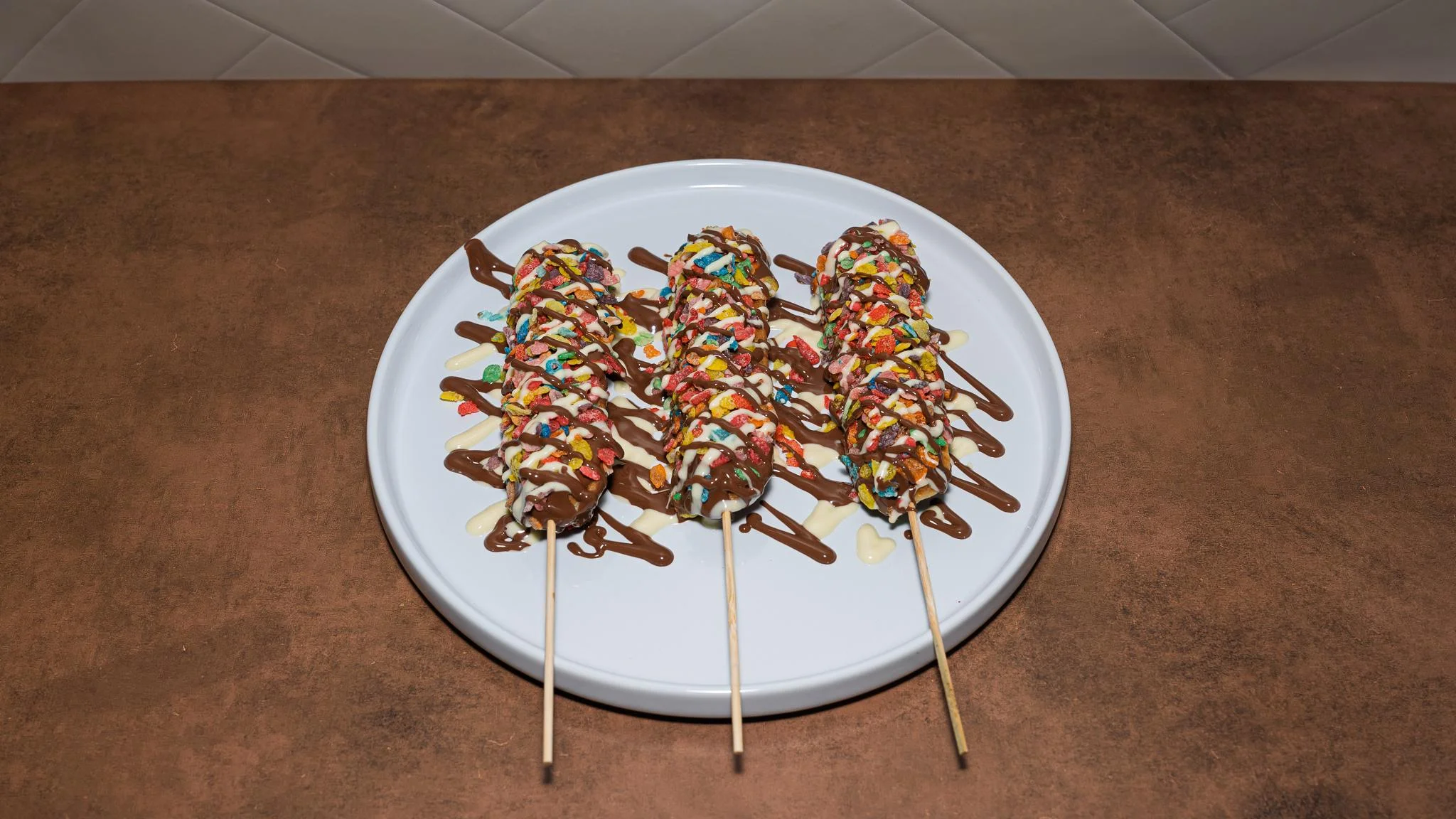 Three dessert skewers coated in chocolate and colorful sprinkles, arranged on a round white plate.