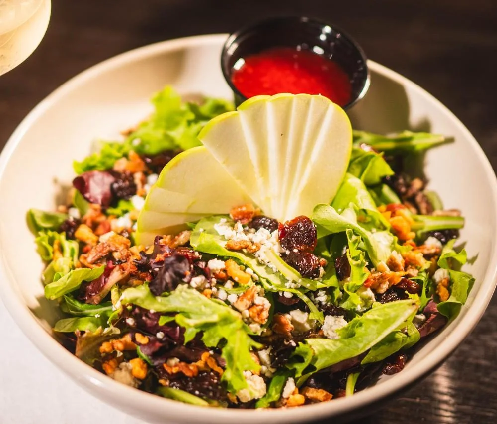 A fresh salad with mixed greens, sliced apples, nuts, and dried cranberries, served with a small bowl of dressing on the side.