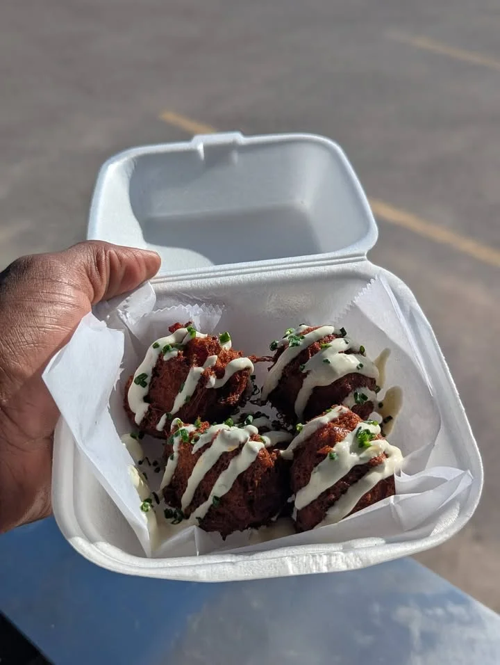 A takeout container holding four round, deep-fried food balls drizzled with sauce and garnished with herbs.
