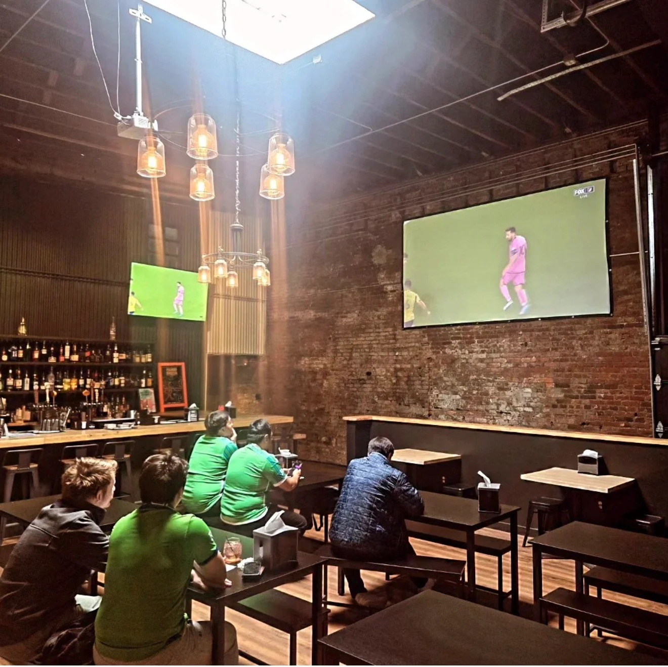 An indoor bar with multiple screens showing a sports game. Patrons are seated at tables, enjoying the atmosphere.