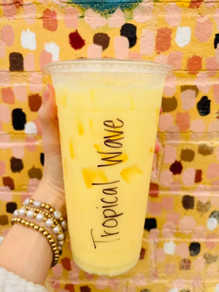 A hand holding a clear cup filled with a bright yellow drink, labeled 'Tropical Wave', against a colorful painted wall.