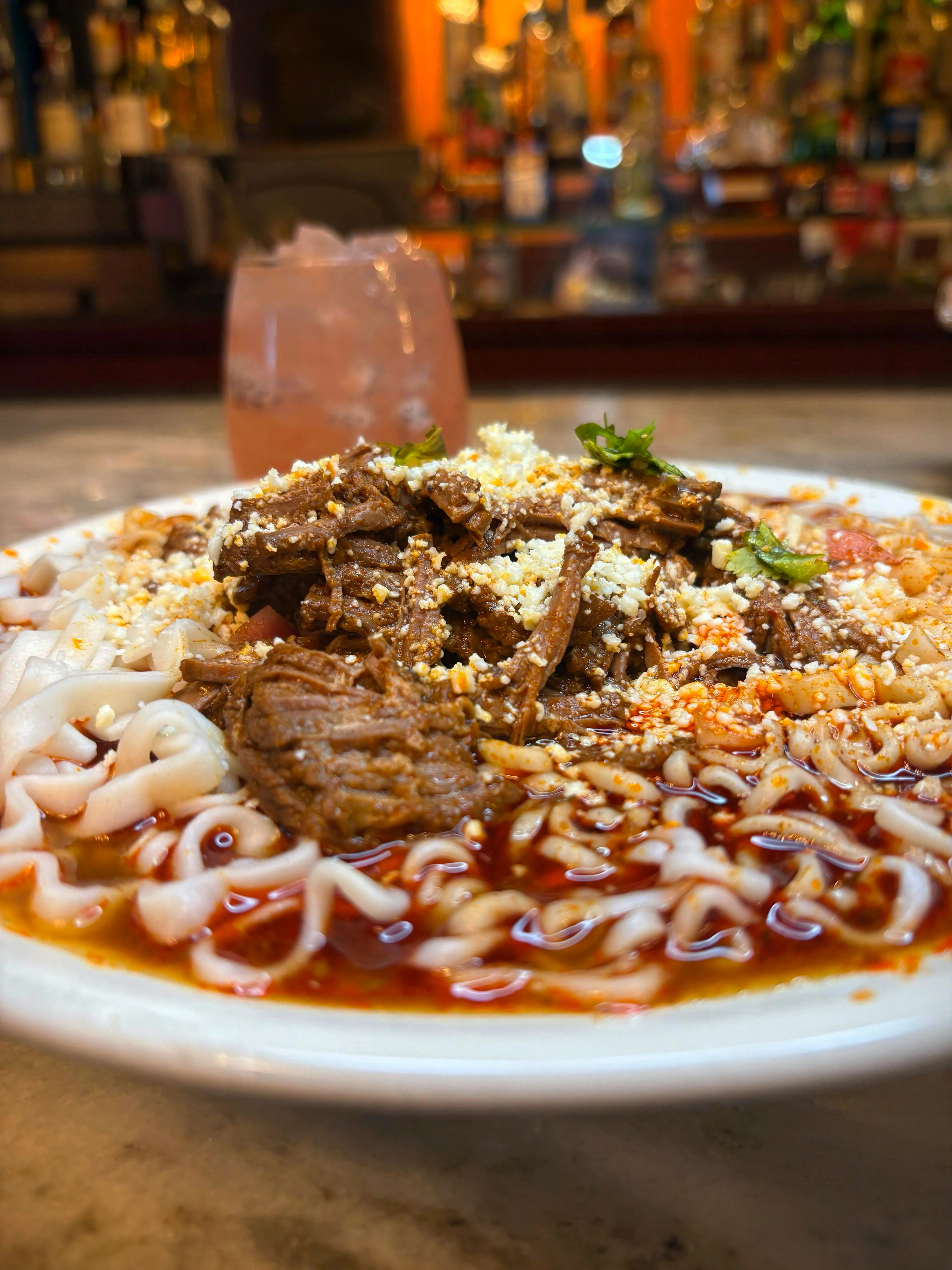 A bowl of noodles topped with shredded beef and sprinkled with cheese and cilantro, served with a drink in the background.