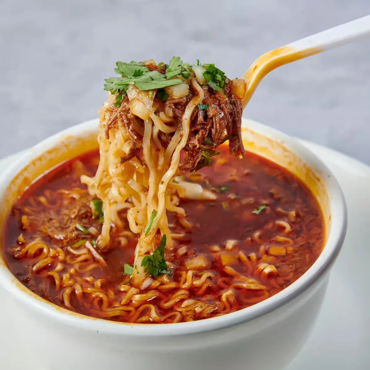 A bowl of spicy noodle soup with tender pieces of meat, garnished with fresh herbs and a fork lifting noodles out of the bowl.