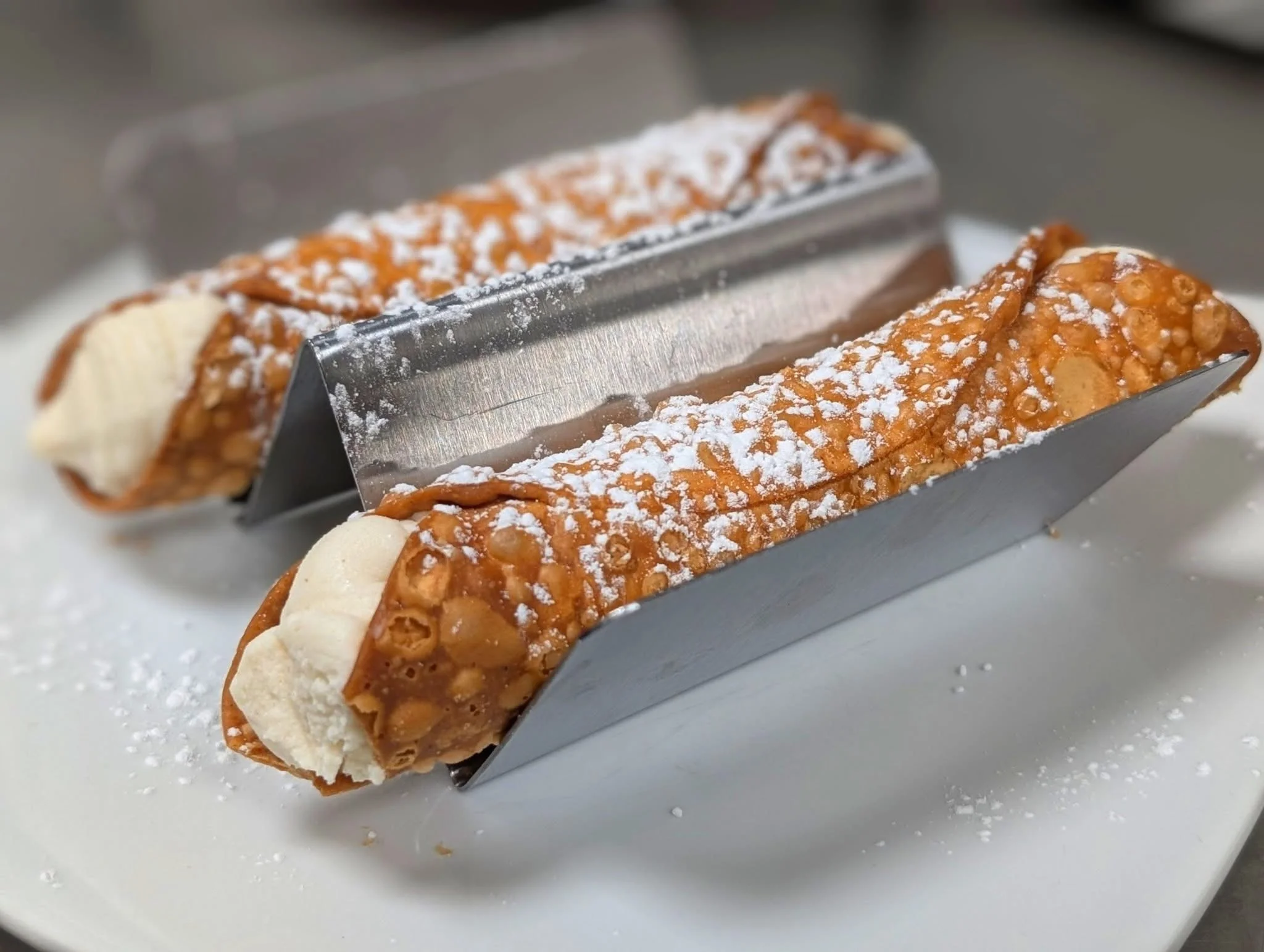Two cannoli filled with creamy filling, presented in metal holders and dusted with powdered sugar, on a white plate.