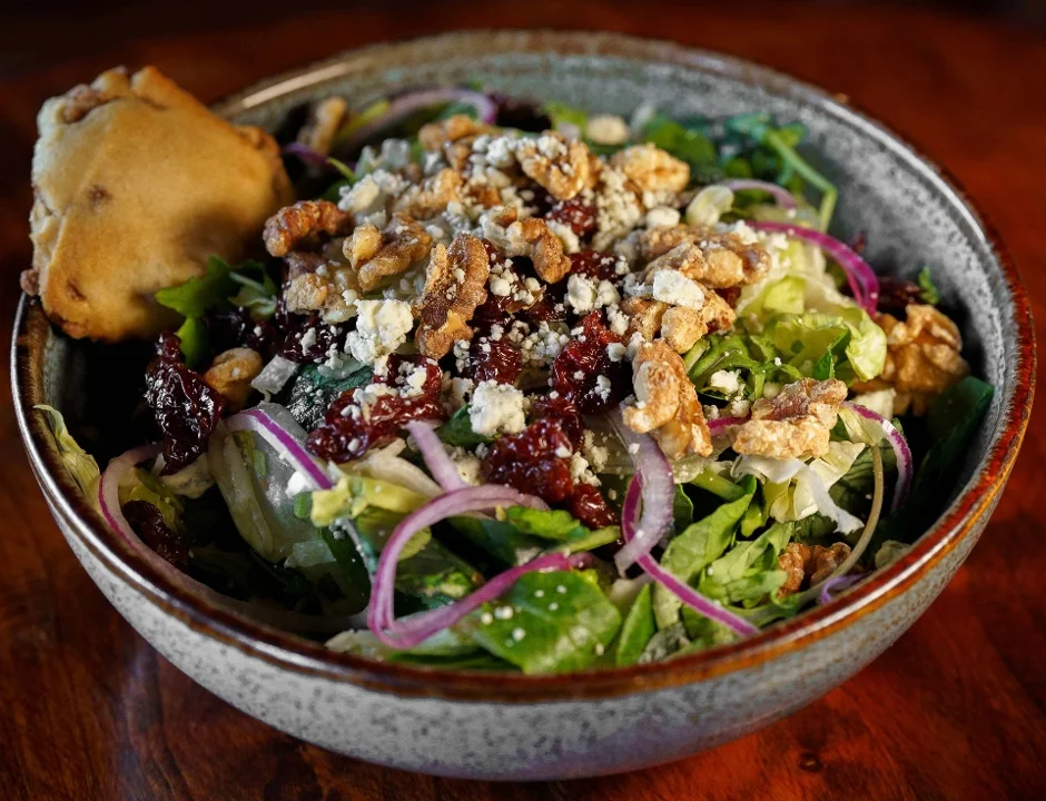 A colorful salad bowl featuring mixed greens, walnuts, cranberries, and red onions, with a small baked good on the side.