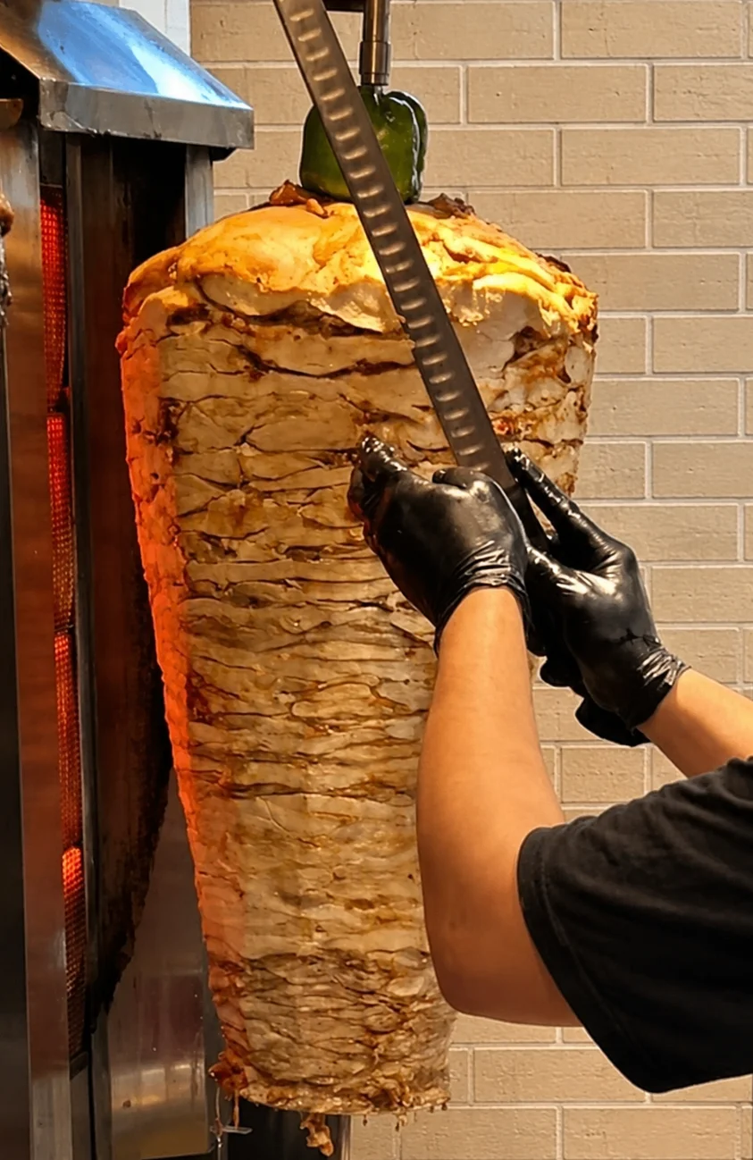 an arab man wearing black gloves cutting slices of chicken shawarma from vertical spit while it is roasting. perfect fresh.