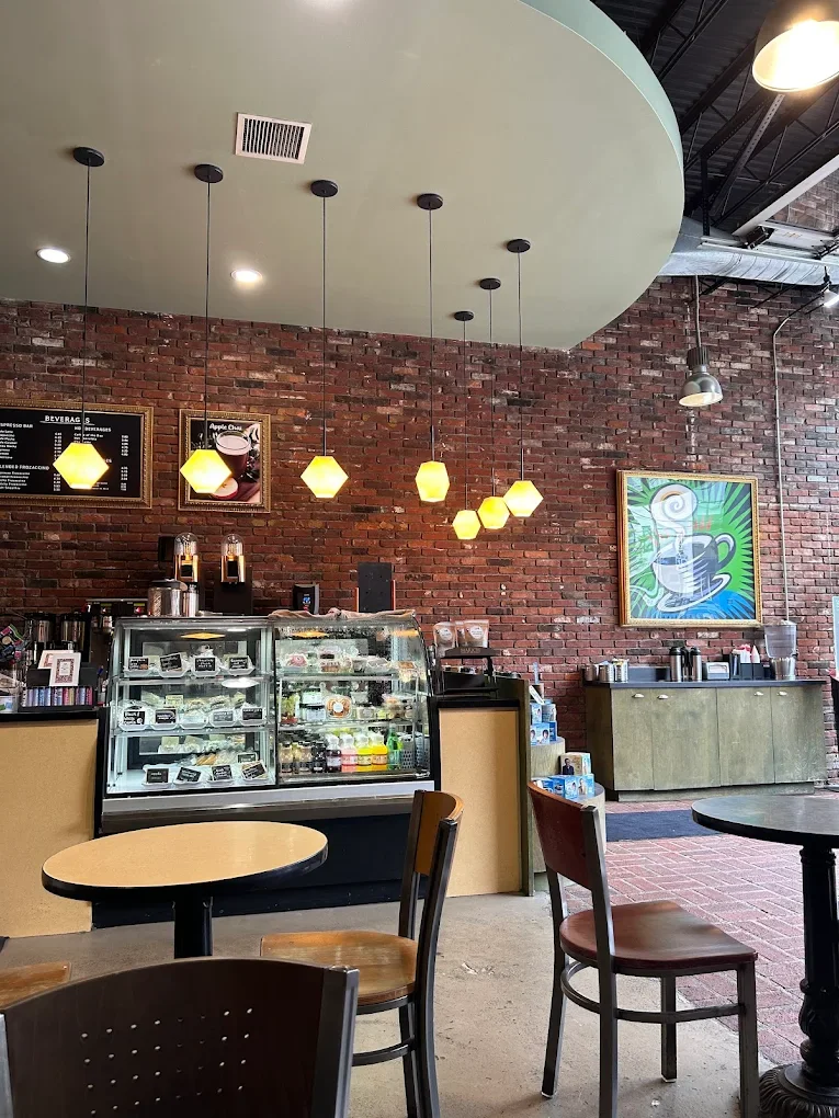 Cozy café interior with exposed brick walls, warm pendant lights, and modern art. Display case with pastries, tables with wooden chairs.