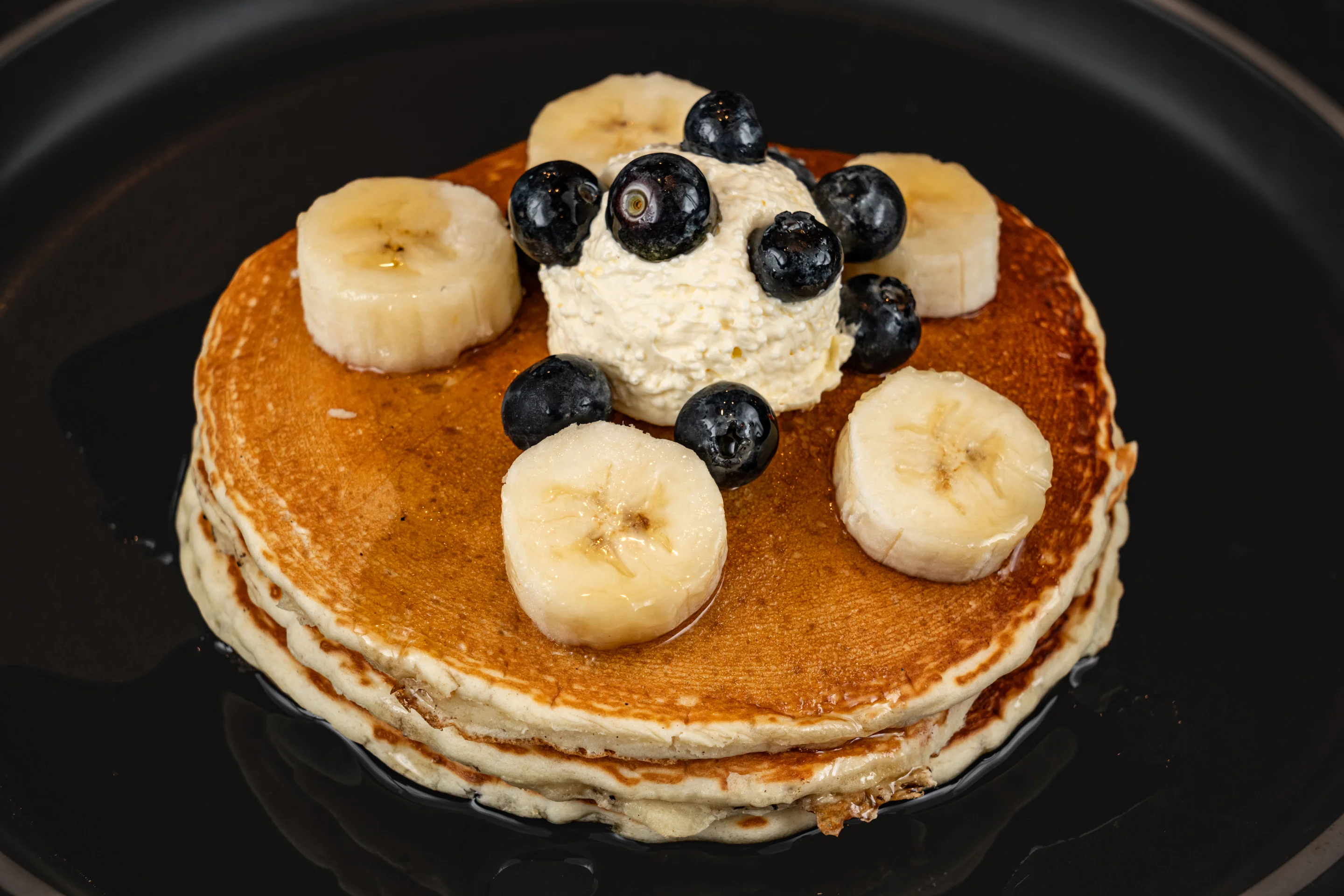 A stack of fluffy pancakes topped with banana slices, blueberries, and a scoop of creamy topping, drizzled with syrup.