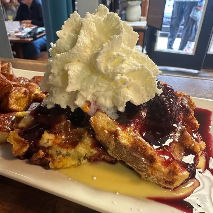 A delicious stack of waffles topped with whipped cream and berry sauce, drizzled with syrup, served on a white plate.