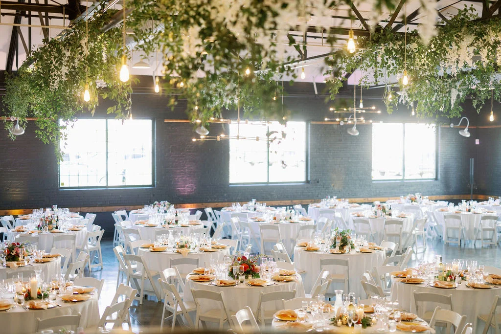 An elegantly arranged banquet space featuring multiple round tables with white tablecloths, glassware, and floral centerpieces. Lush greenery adorns the ceiling, enhancing the ambiance.