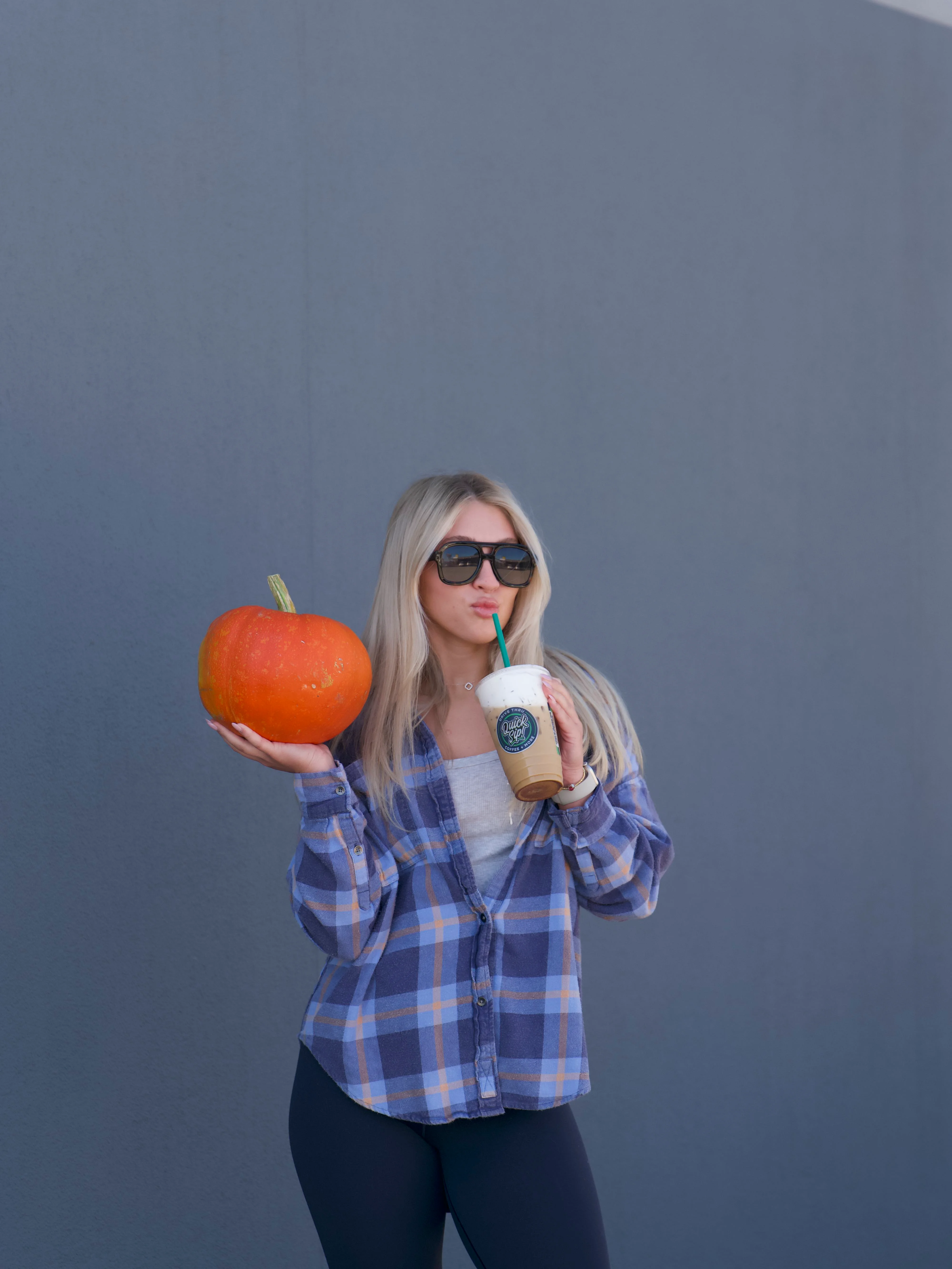 Woman holding a pumpkin and iced coffee against a gray wall