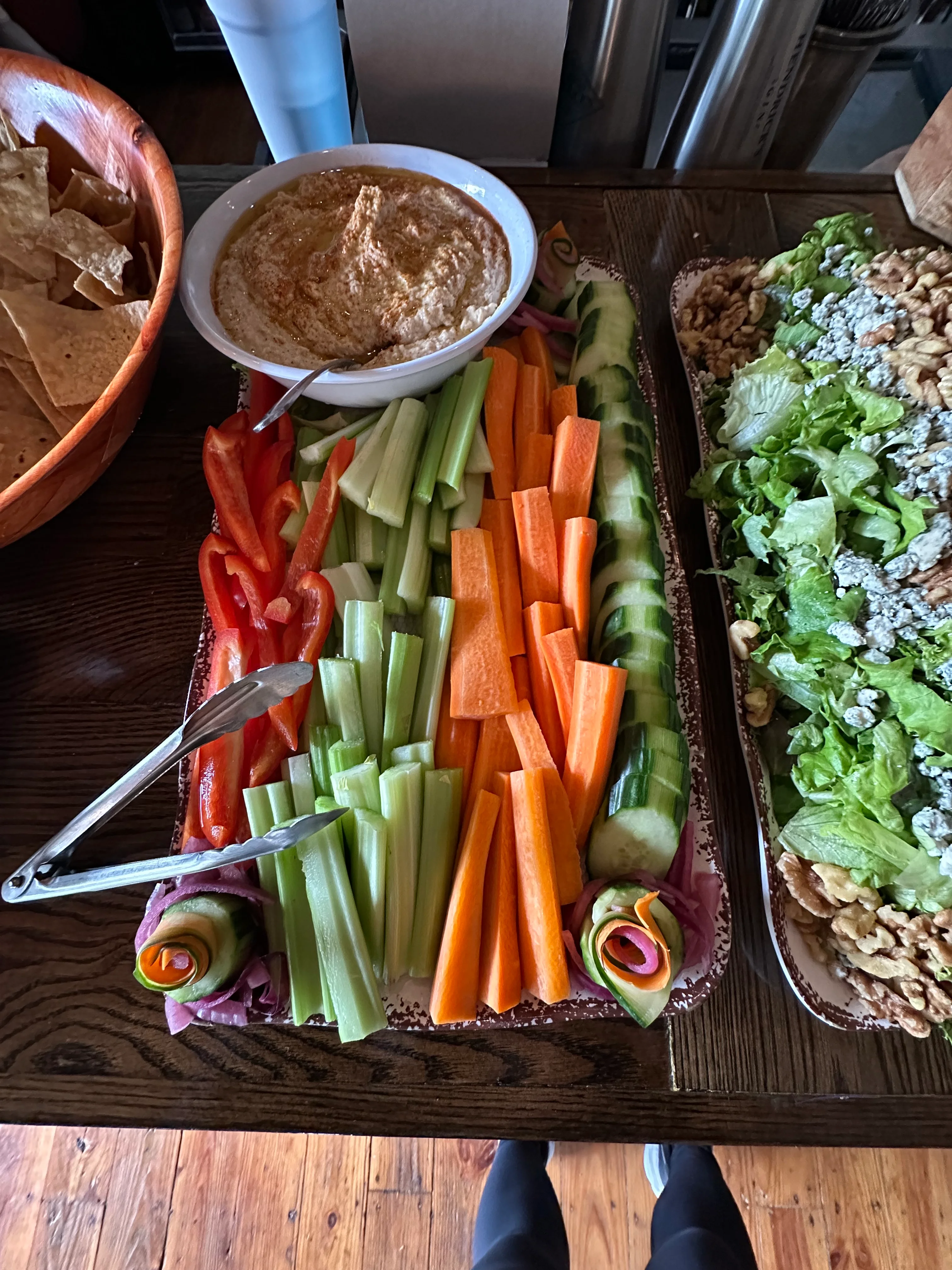 A colorful vegetable platter featuring red peppers, cucumbers, carrots, and celery, served with a bowl of hummus. Tongs are placed nearby for serving.