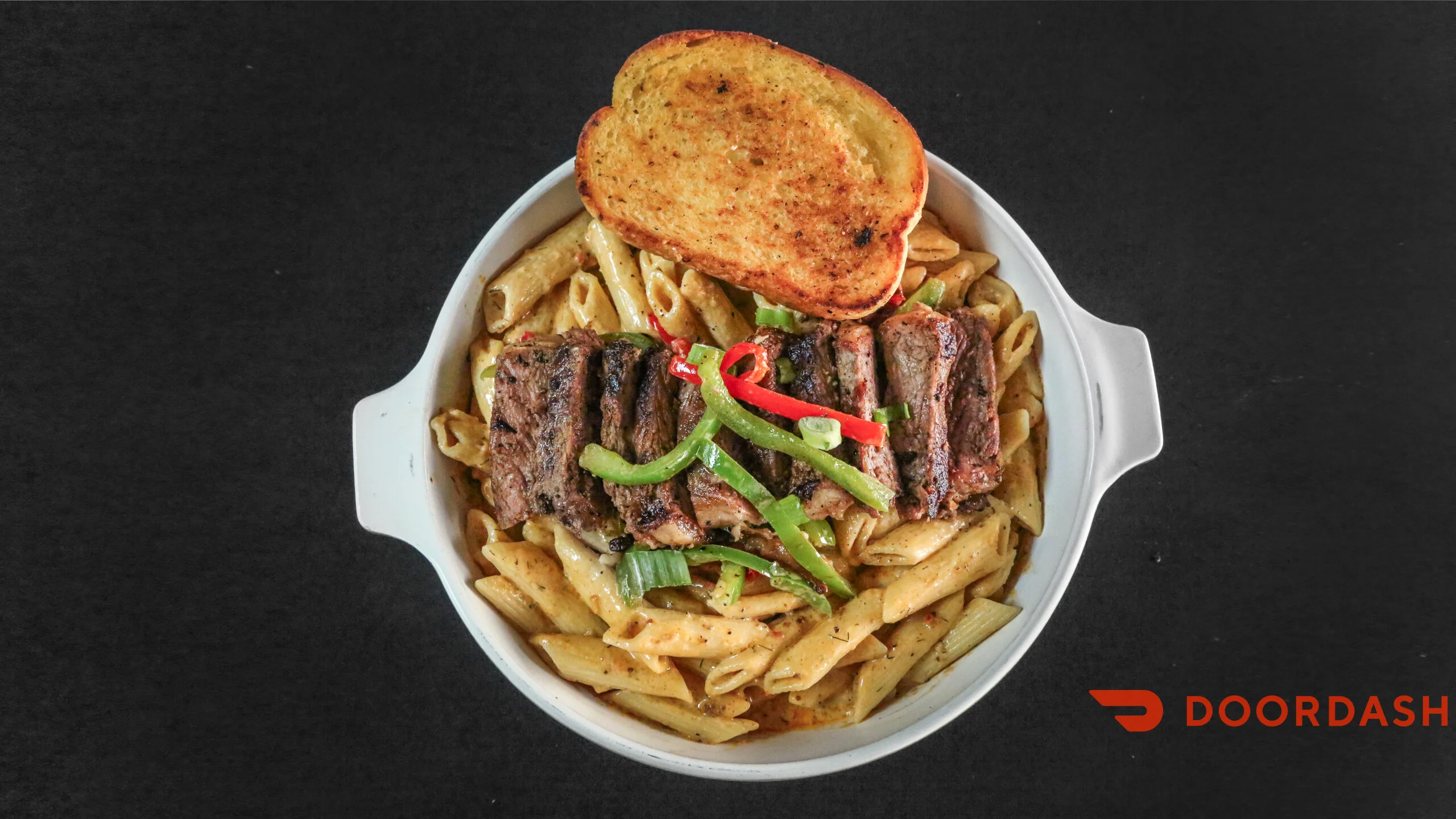 A bowl of pasta topped with grilled steak, green peppers, and slices of toasted bread. The dish features penne pasta in a creamy sauce with colorful toppings.