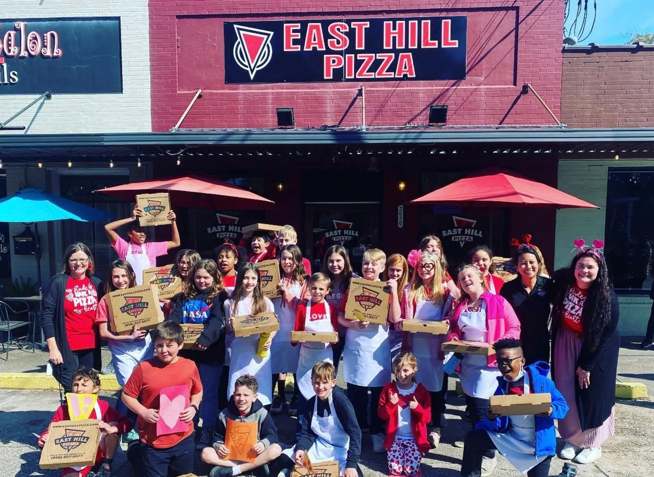 Community group posing outside a neighborhood pizzeria with pizza boxes