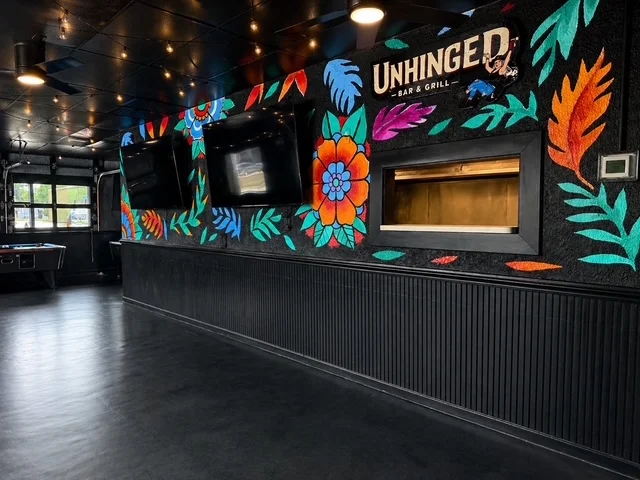 A modern bar and grill interior featuring vibrant floral wall art. The space is accentuated with dark elements and glowing lights.