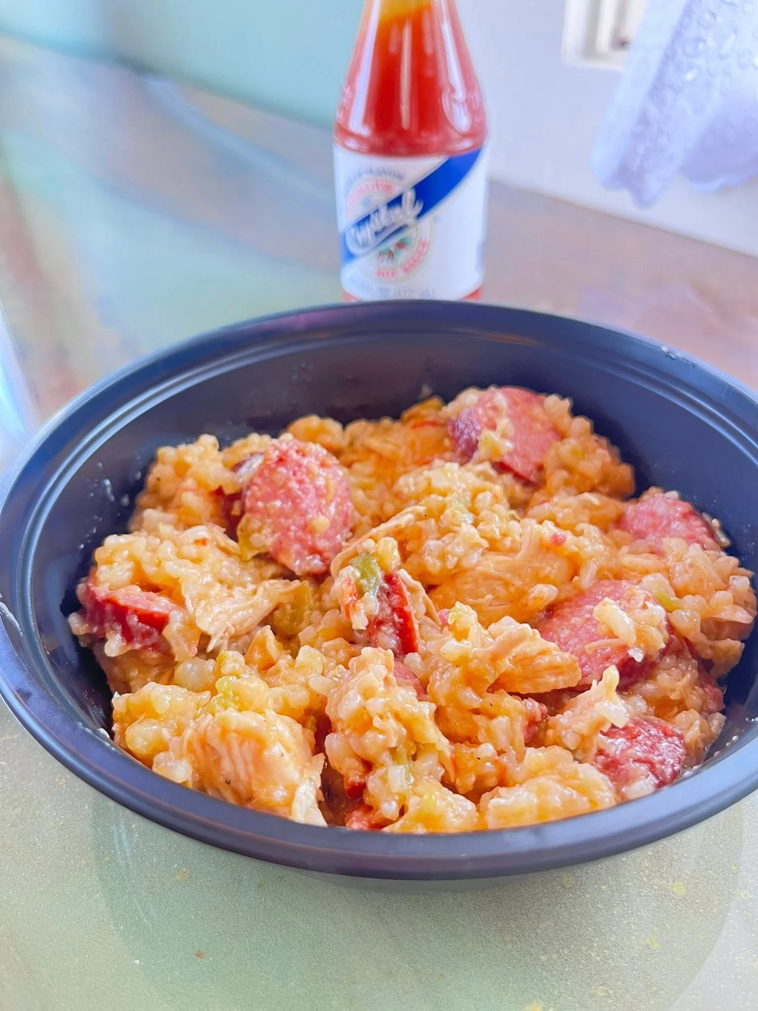 Bowl of rice mixed with chicken and sausage, with hot sauce in the background.
