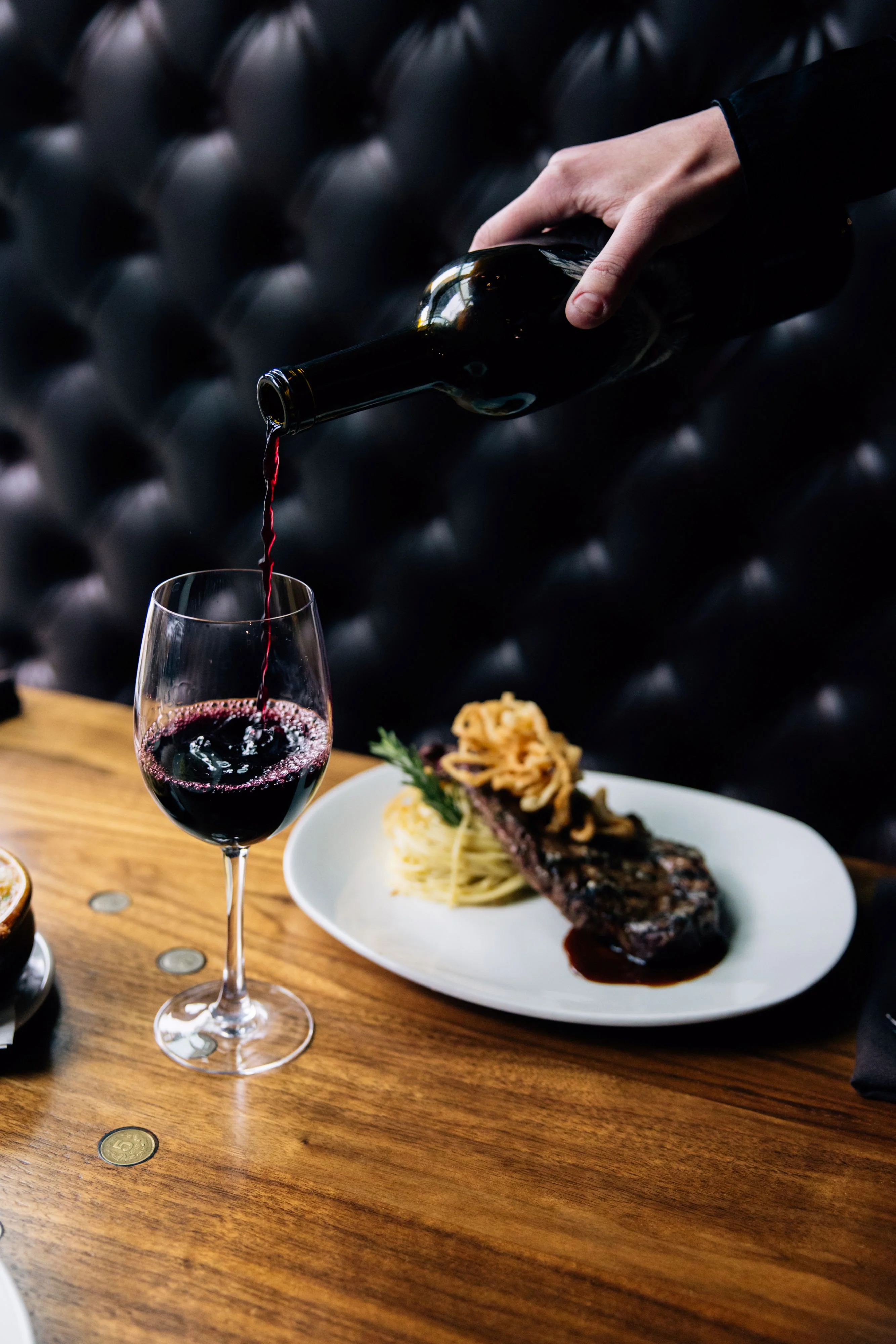 Red wine being poured into a glass beside a steak and pasta dish.