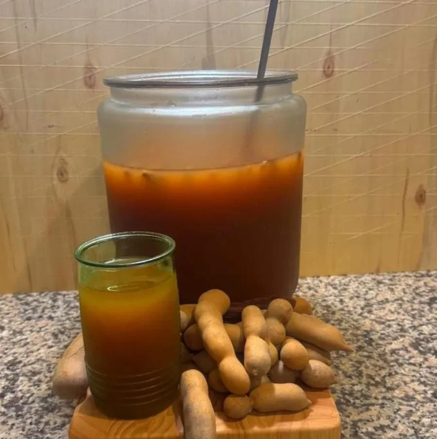 A jar of tamarind drink with a glass and tamarind pods placed on a wooden board.