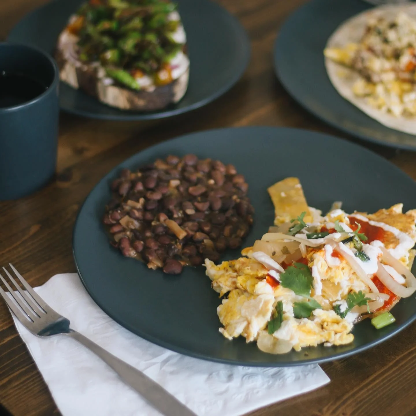 A plate of breakfast eggs with black beans, accompanied by toast topped with vegetables, and a dark-colored beverage in a cup.