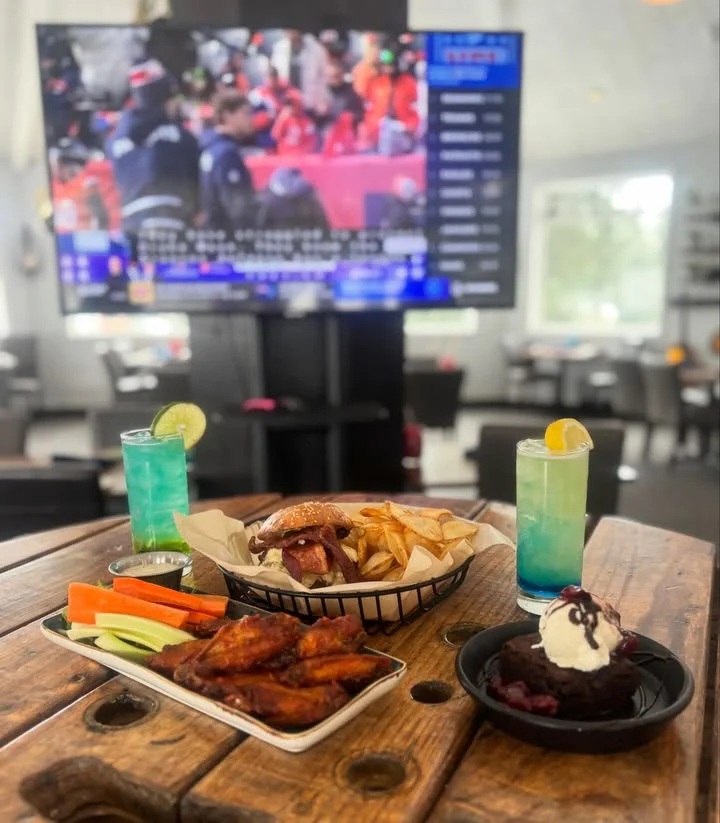 A table spread with a burger, fries, chicken wings, vegetable sticks, and a rich chocolate dessert, alongside colorful drinks and a sports broadcast in the background.