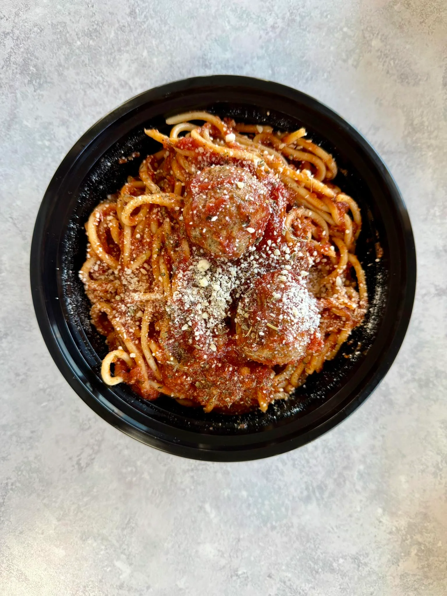 A black bowl filled with spaghetti topped with marinara sauce and meatballs. The dish is garnished with shredded cheese and herbs.