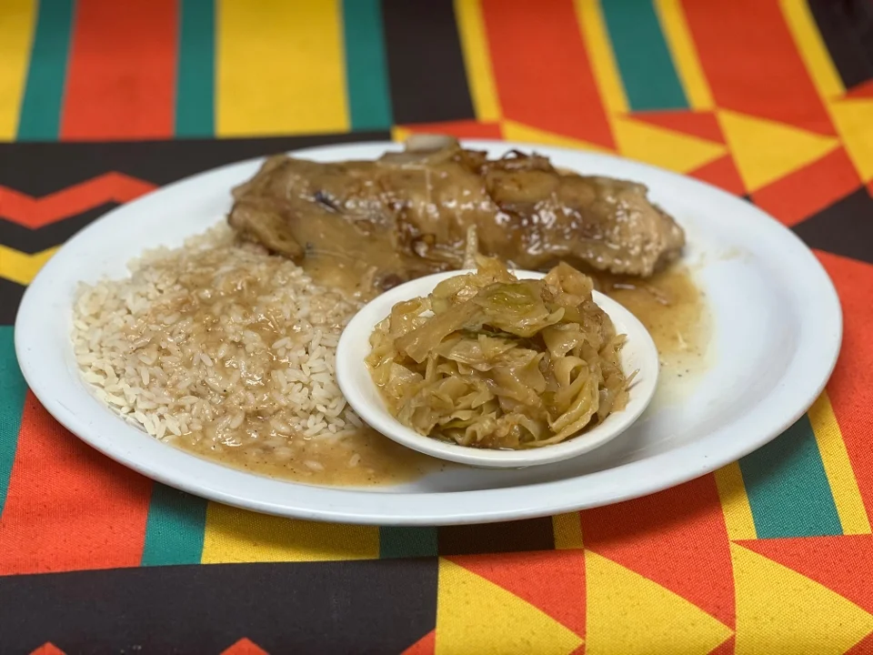 A plate featuring a serving of rice, a piece of fish in gravy, and a side of sautéed cabbage, all presented on a colorful patterned table cover.