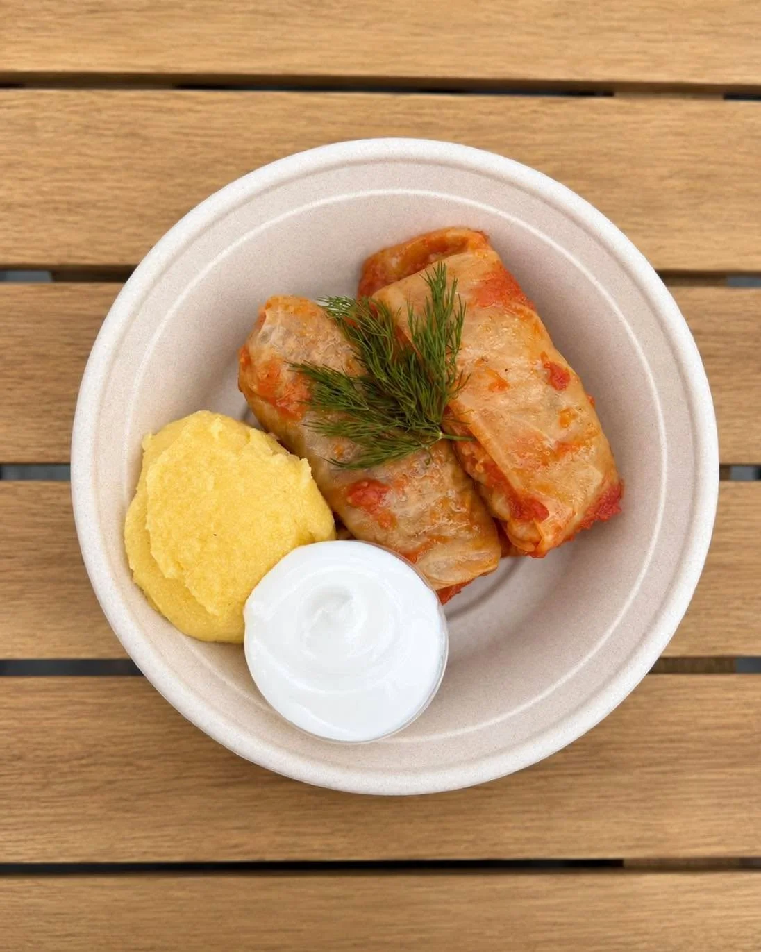 A bowl containing stuffed cabbage rolls served with a side of creamy polenta and sour cream, garnished with fresh dill.