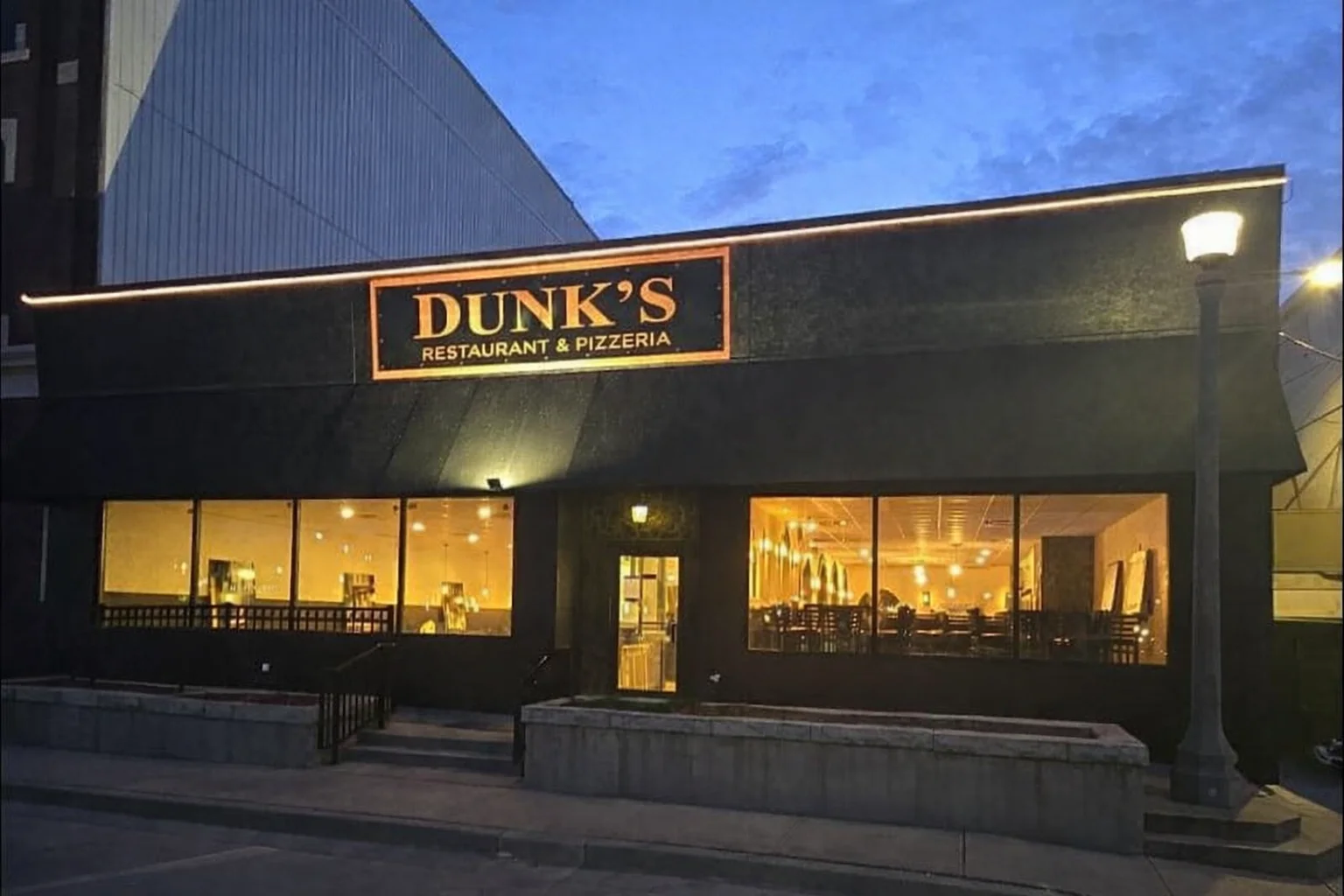An exterior view of a restaurant and pizzeria illuminated in the evening, featuring large windows that show the interior dining area.