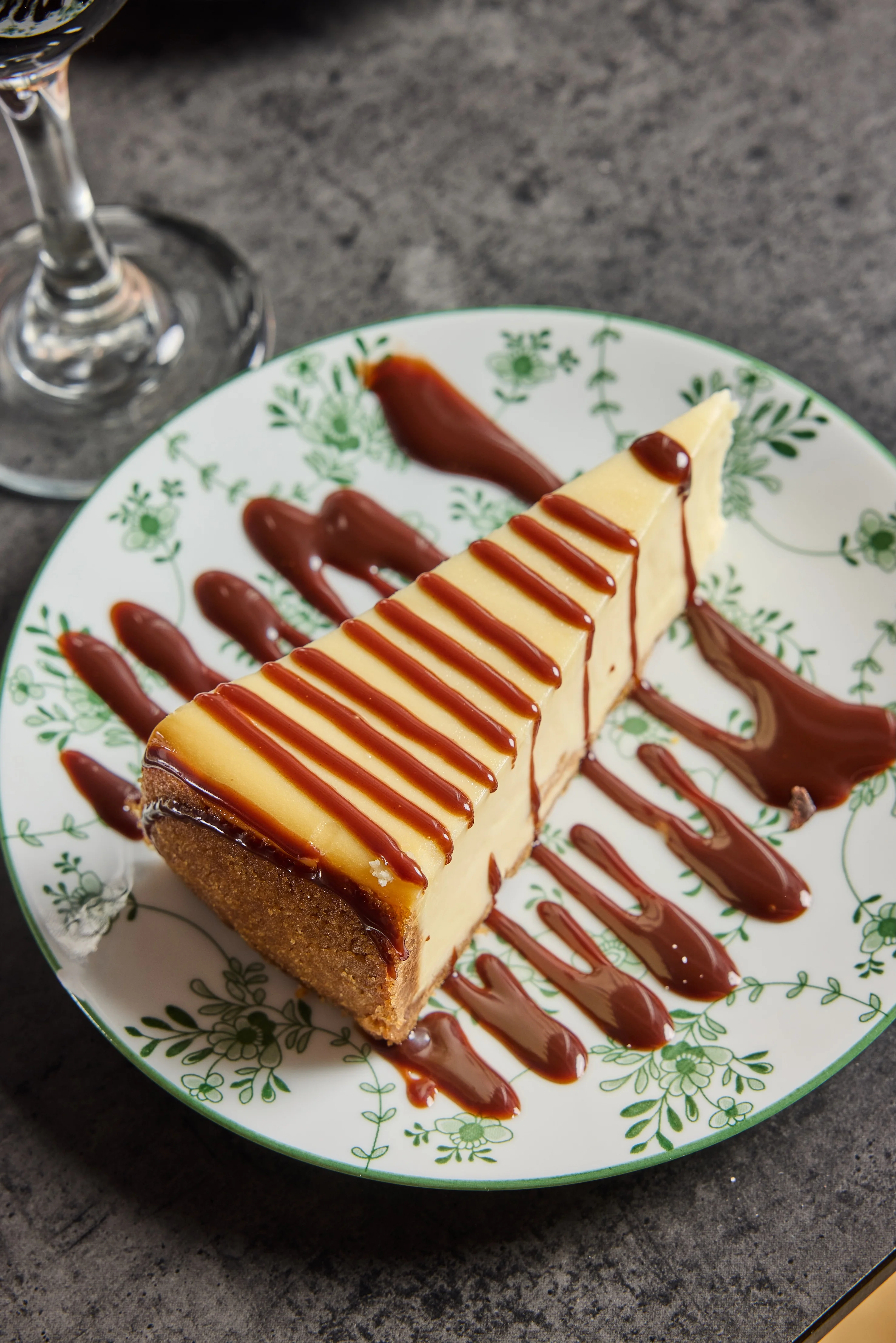 A slice of creamy cheesecake drizzled with chocolate sauce on a floral-patterned plate. The setting is cozy, hinting at indulgence and satisfaction.